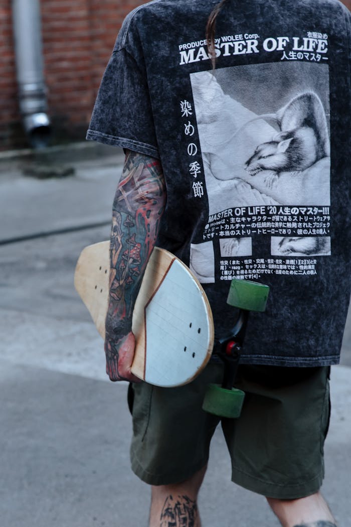 Back view of tattooed skateboarder holding skateboard on urban street. Casual attire and detailed tattoos.